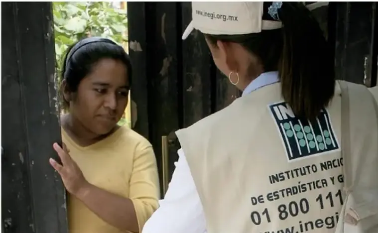 Fotografía de una encuestadora del Inegi con gorra y chaleco que dice 'INEGI' y 'INSTITUTO NACIONAL DE ESTADÍSTICA Y GEOGRAFÍA', hablando con una mujer en la puerta de su casa.