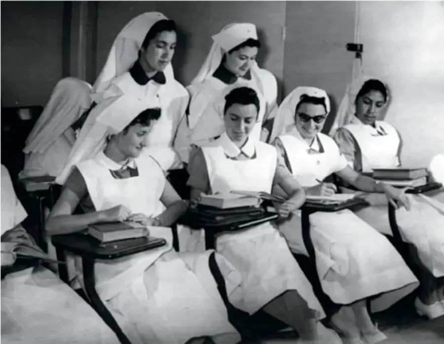 Fotografía en blanco y negro de un grupo de enfermeras o estudiantes de medicina con uniforme blanco y cofia, sentadas en un aula tomando apuntes.