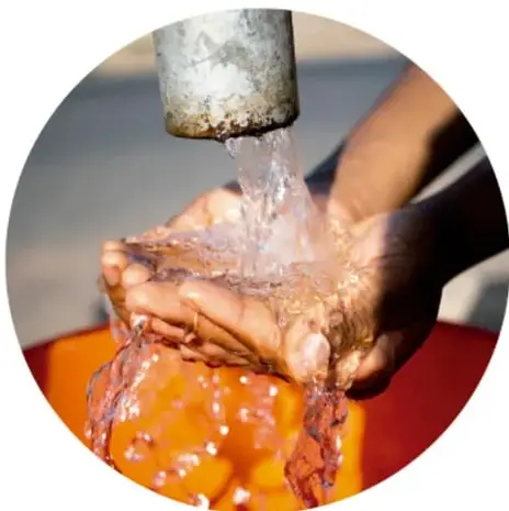 Fotografía de una persona que lava sus manos bajo el chorro de agua que sale de un tubo, con agua espumosa cayendo a un recipiente anaranjado.
