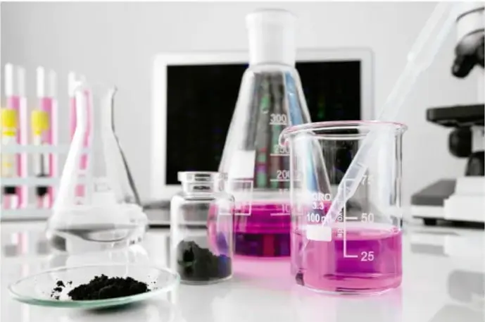 Fotografía de un laboratorio con matraces, vasos con líquido rosado, portaobjetos con sustancia negra y microscopio.