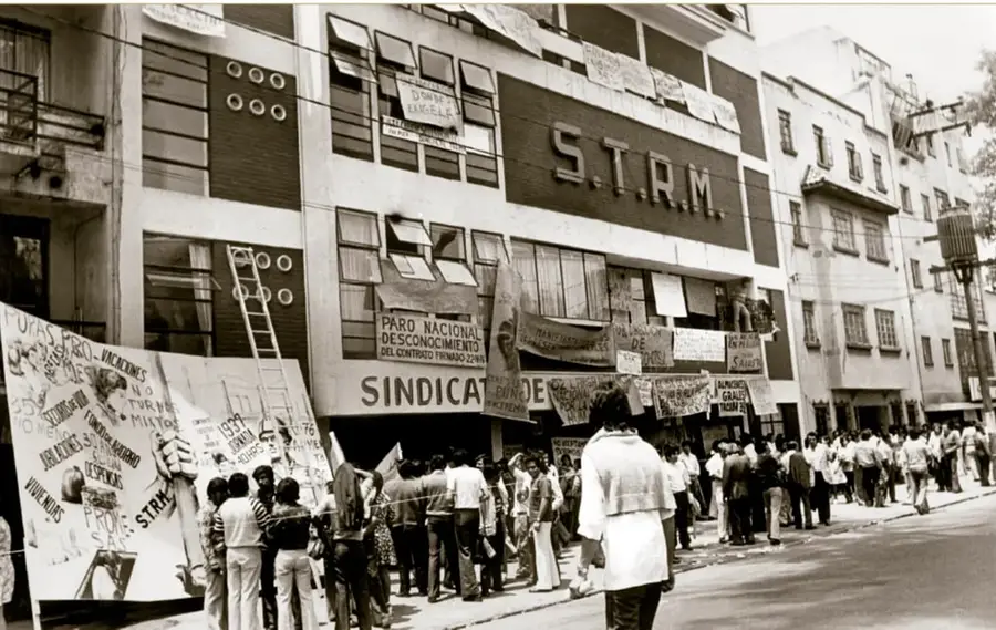 Fotografía en blanco y negro del frente del edificio del Sindicato de Telefonistas (S.T.R.M.) con letreros de 'SINDICATO' y 'PARO NACIONAL DESCONOCIMIENTO DEL CONTRATO FIRMADO', múltiples cartelas con peticiones (vacaciones, aumento de sueldo, descanso semanal) y una multitud de trabajadores en la banqueta.
