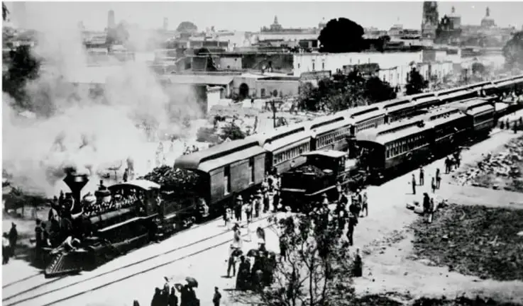Fotografía en blanco y negro de una locomotora de vapor con varios vagones y personas alrededor, en un paisaje urbano con edificios al fondo. Representa la llegada del ferrocarril durante el Porfiriato.