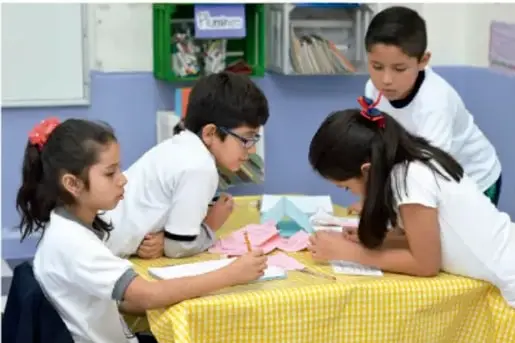 Fotografía de cuatro estudiantes de primaria trabajando en equipo sobre una mesa con mantel amarillo.