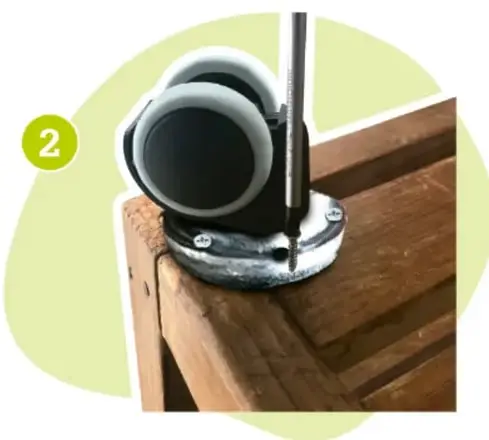 Primer plano de una rueda negra con placa metálica siendo atornillada a la esquina de madera del huacal con un desarmador.