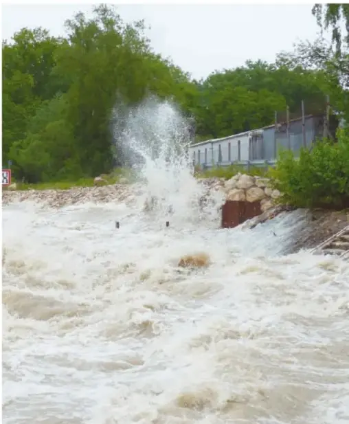 Corriente de agua turbia y crecida que salpica junto a árboles y una estructura metálica, señal de inundación.