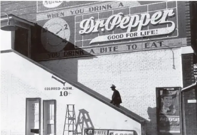 Fotografía histórica en blanco y negro que muestra un edificio con un anuncio publicitario de la bebida Dr Pepper. En la parte inferior se observa un letrero que dice 'COLORED ADM 10c', indicando una entrada segregada para personas afrodescendientes, con una escalera y una persona subiendo. Refleja la segregación racial en espacios públicos.