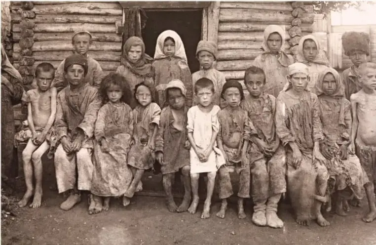 Fotografía histórica en blanco y negro de un grupo de campesinos rusos, en su mayoría niños y mujeres con ropa raída, sentados y de pie frente a una vivienda de troncos de madera. Refleja la pobreza del campesinado ruso a inicios del siglo XX.