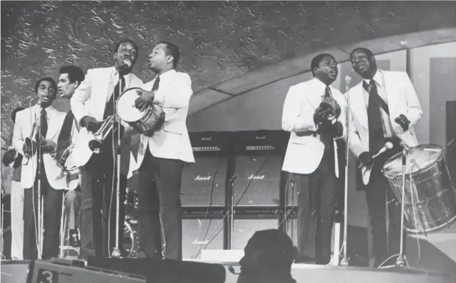 Fotografía en blanco y negro de un grupo de músicos con trajes claros tocando tambores y cantando en un escenario.