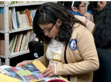 Fotografía de una estudiante con uniforme revisando materiales impresos en una biblioteca escolar.