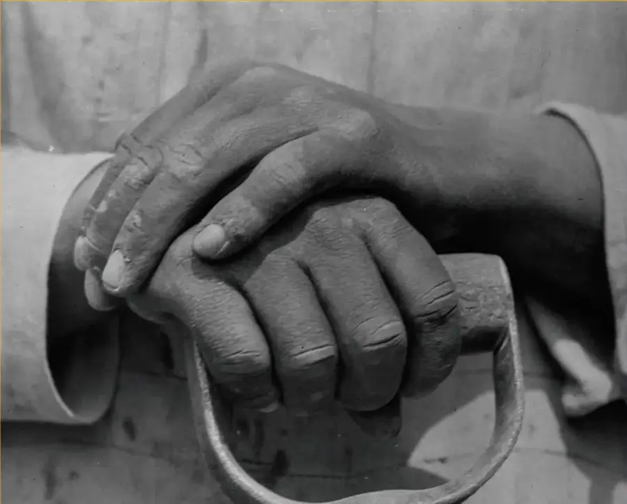 Fotografía en blanco y negro de dos manos de diferentes tonos entrelazadas sobre el mango metálico de una herramienta, símbolo de colaboración humana y comunitaria.