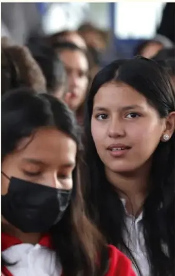 Estudiantes de secundaria en una asamblea; una joven con cubrebocas y otra al frente, atentas a la reunión.