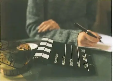 Fotografía de manos de una persona escribiendo en papel con bolígrafo. Al fondo se ven clappers (claquetas) de filmación cinematográfica, evocando el contexto de la reseña de películas. Crédito: Shutterstock (2020) 540260491.
