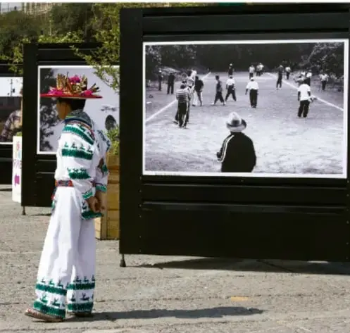 Fotografía de una persona con vestimenta tradicional de colores vivos frente a un gran muro o pantalla urbana que muestra una imagen en blanco y negro de una escena con varias personas en un espacio público; representa el arte de gran formato en el espacio urbano.