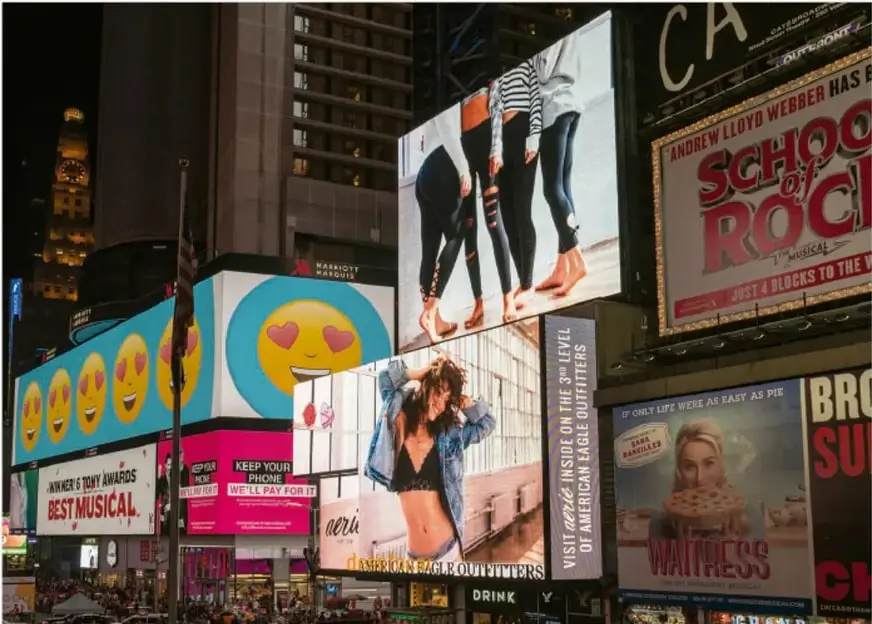 Fotografía de un paisaje urbano nocturno con múltiples anuncios espectaculares luminosos, entre ellos uno con emojis sonrientes, otro de una marca de mezclilla y un cartel del musical "School of Rock".