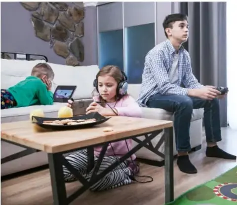 Fotografía de tres adolescentes en una sala de estar: uno acostado con tableta, una niña con audífonos y un joven con control, en actividad de convivencia.