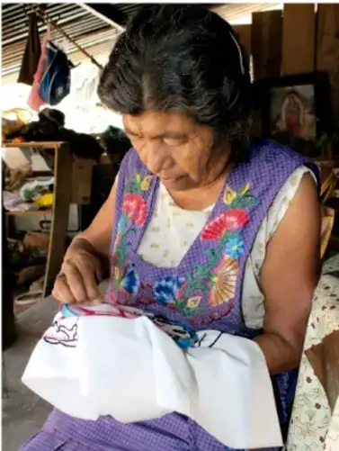 Fotografía de una artesana indígena mayor bordando una tela blanca con hilos de colores, sentada en un taller comunitario; ejemplifica la creación artística comunitaria y la inclusión cultural.