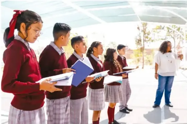 Grupo de estudiantes de telesecundaria en uniforme (suéteres rojos y faldas/pantalones gris a cuadros) sosteniendo carpetas y carpetas de trabajo, junto a su maestra, en un espacio escolar abierto; al parecer ensayan o reciben indicaciones para la presentación de su proyecto.