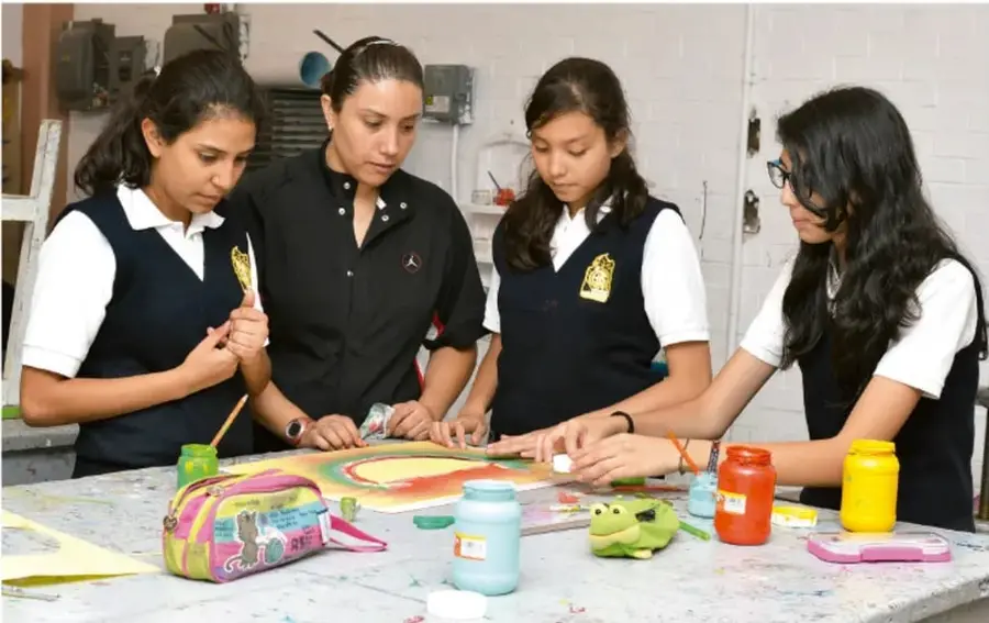 Fotografía de cuatro estudiantes y una docente trabajando juntas en una mesa con pinturas, pinceles y materiales de arte, realizando una actividad creativa colectiva.