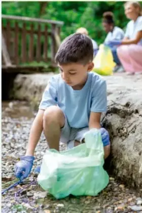 Fotografía de un niño recolectando basura en una bolsa en un espacio natural con adultos al fondo.