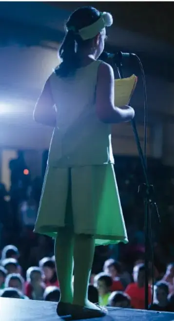 Fotografía de una niña o joven sobre un escenario, iluminada con luz verde, sosteniendo un papel y frente a un micrófono; al fondo se distingue un público sentado. Ilustra la tradición oral y la recitación de textos.