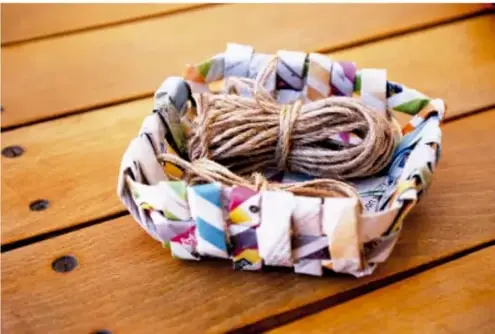 Canasta elaborada con tiras de papel de colores tejidas, con un rollo de hilo de cáñamo en su interior, colocada sobre una mesa de madera con vetas visibles.