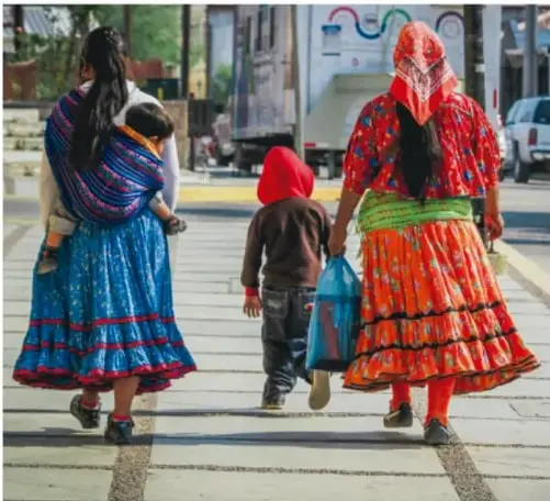 Fotografía de dos mujeres y una niña ralámuli caminando por una calle: faldas amplias de colores (azul y rojo-naranja), pañuelos en la cabeza; una carga a un bebé.