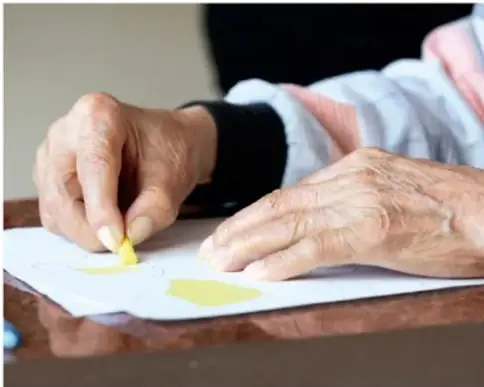 Manos de una persona mayor coloreando con un lápiz pastel amarillo sobre una hoja, ejemplo de actividad de arte-terapia.