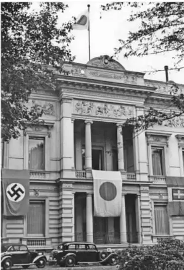 Fotografía histórica en blanco y negro de un edificio gubernamental donde ondean una bandera con la esvástica nazi y una bandera japonesa; al frente hay automóviles de época, ilustrando la sede de una potencia del Eje.
