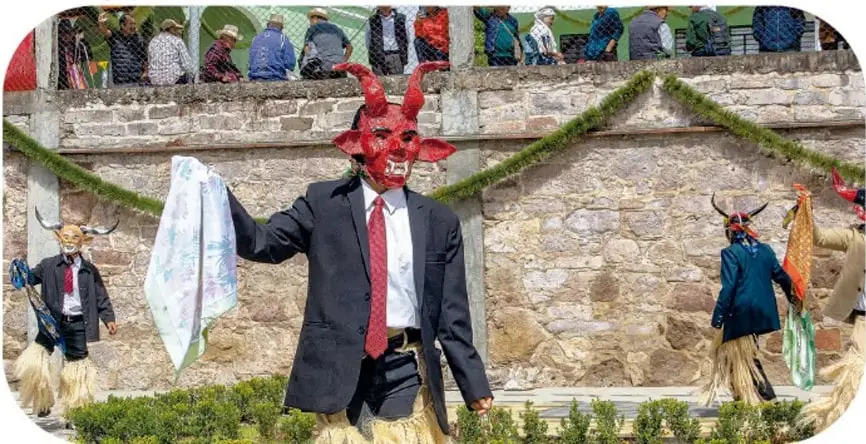 Fotografía de danzantes del baile de los diablos: un hombre con máscara roja de cuernos, saco negro, camisa blanca y corbata roja sostiene un pañuelo blanco; otros danzantes con máscaras y faldas de fibra bailan frente a un muro de piedra, con público observando desde arriba. Crédito: Jorge López Vela, Ciudad de México.
