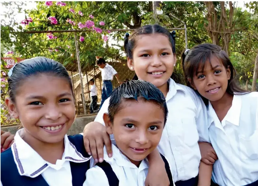 Fotografía de cuatro estudiantes de primaria con uniforme escolar sonriendo al aire libre, con árboles y flores de fondo.