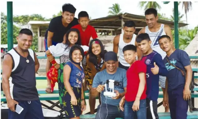Fotografía de un grupo de jóvenes y adolescentes, algunos sentados y otros de pie, sonriendo en un espacio abierto con estructura de madera y vegetación al fondo. Parecen ser estudiantes de secundaria o bachillerato.