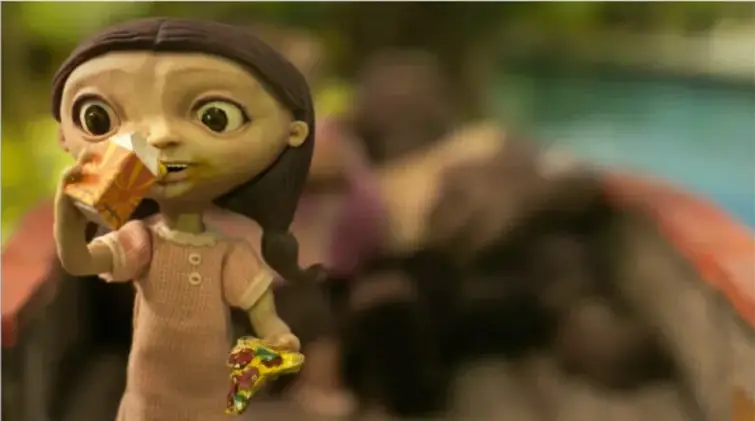 Fotograma del cortometraje: una niña con ojos grandes y cabello trenzado come una rebanada de pizza mientras sostiene un vaso; al fondo se ve un parque acuático desenfocado.