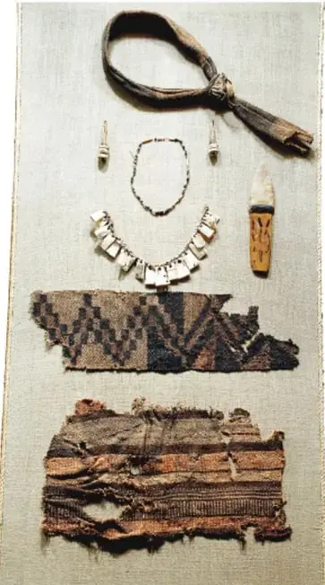 Fotografía de artefactos prehispánicos: un cinturón de cuero con nudo, collares de cuentas y piedras, un cuchillo o instrumento con mango decorado, y fragmentos de textiles bordados con motivos geométricos, sobre fondo claro.