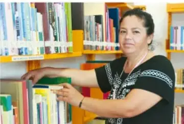Fotografía de una mujer organizando libros en una estantería de biblioteca o aula, en tonos naranjas y verdes.