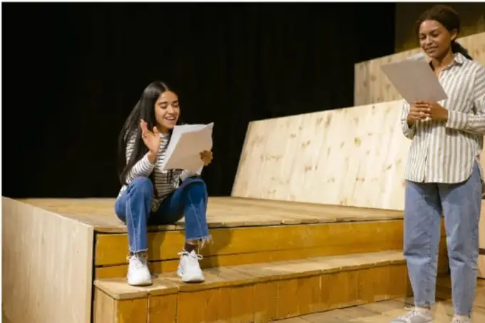Fotografía de dos estudiantes en un escenario de madera con gradas; una sentada leyendo un documento y otra de pie sosteniendo una hoja, aparentemente ensayando o exponiendo los resultados de su proyecto.