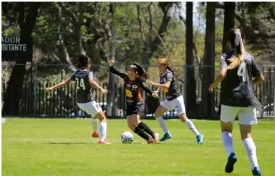 Fotografía de un partido de fútbol femenil en cancha de pasto con jugadoras de dos equipos disputando el balón, árbitra y espectadores al fondo entre árboles.