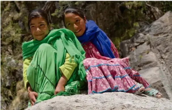 Fotografía de dos niñas indígenas con rebozos de colores verde y azul, sentadas en un entorno rocoso de montaña.