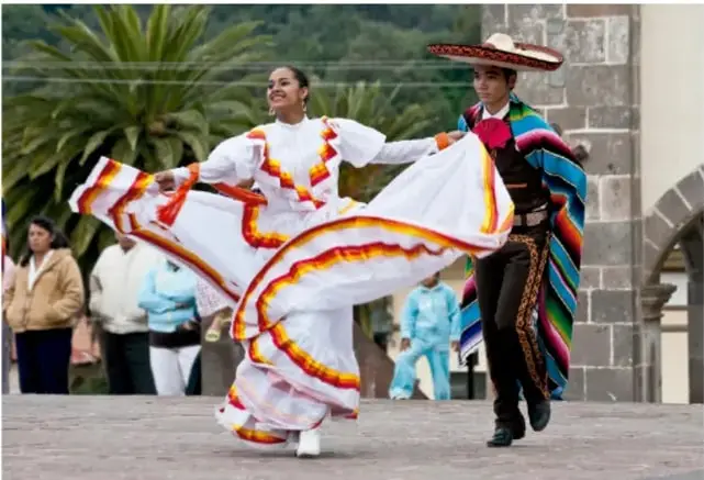 Pareja bailando danza folclórica mexicana al aire libre: mujer con vestido blanco de volantes con franjas naranjas y rojas, hombre con sombrero grande y sarape de colores; al fondo, gente observando y un edificio de piedra.