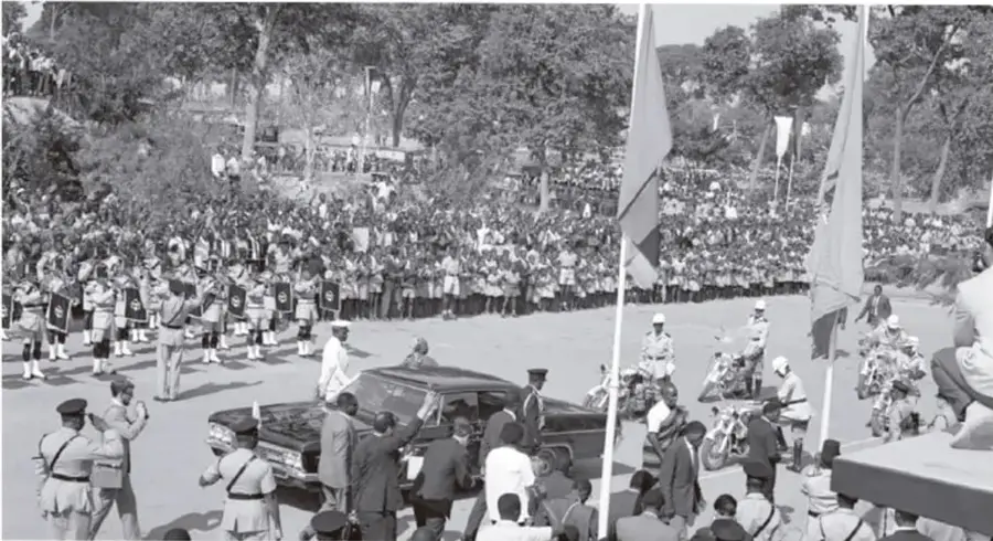 Fotografía en blanco y negro de una multitud reunida en una plaza con banderas, vehículos oficiales y personas uniformadas; parece un acto público o desfile relacionado con la transición democrática sudafricana.