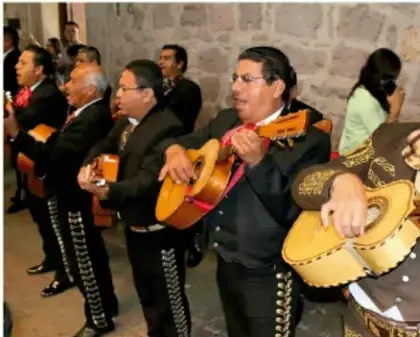 Fotografía de un grupo de músicos vistiendo trajes tradicionales (chaquetas bordadas) y tocando guitarras y guitarrones en un espacio con muros de piedra, ilustrando un rasgo cultural de una comunidad.