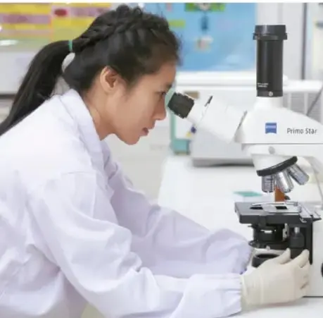 Fotografía de una estudiante con bata blanca observando a través de un microscopio en un laboratorio escolar.