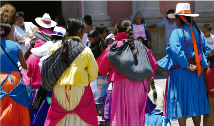 Fotografía de mujeres indígenas de espaldas con vestidos tradicionales de colores vivos (azul, amarillo, rosa) y sombreros, cargando rebozos y bolsas, en un espacio público con público alrededor.