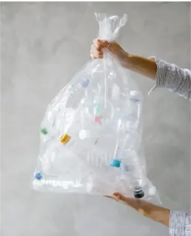 Manos sosteniendo una bolsa transparente llena de botellas de plástico de colores, sobre fondo de concreto.