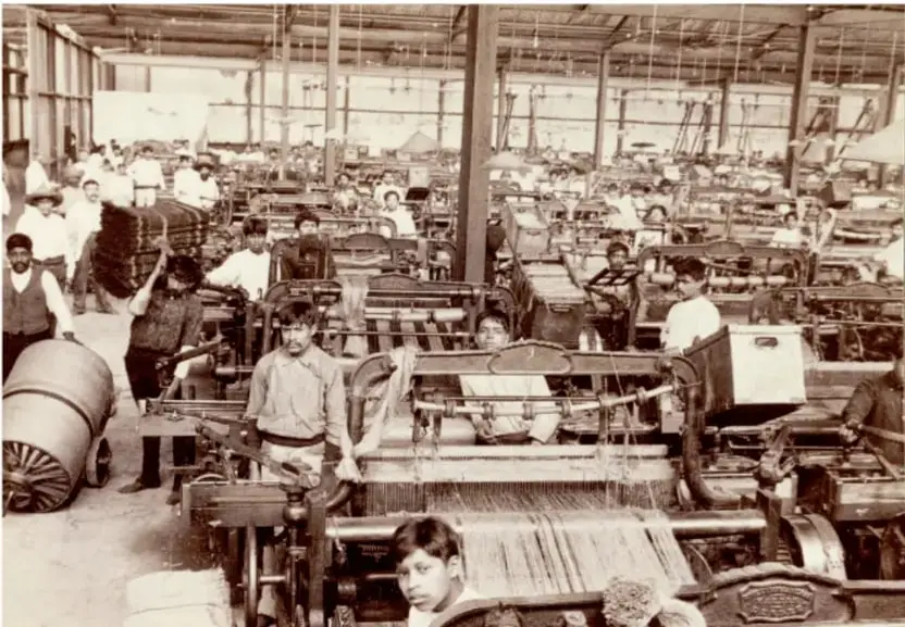 Fotografía en blanco y negro de una fábrica textil de principios del siglo XX. Se observan numerosos obreros, incluidos niños, operando telares mecánicos en un amplio galpón industrial. Refleja la industrialización porfirista y las condiciones laborales de la época.