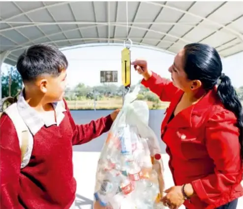 Estudiante y maestra pesando una bolsa de residuos con una balanza de mano, en un espacio techado al aire libre.
