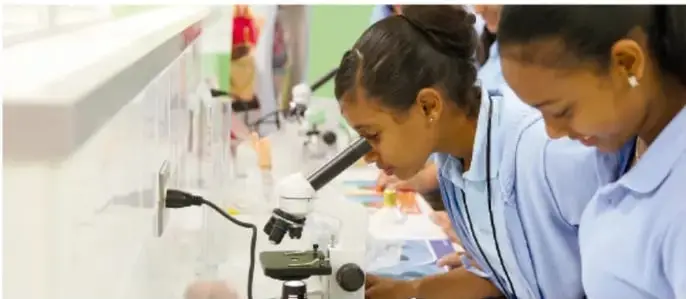 Fotografía de dos niñas con uniforme azul observando por un microscopio en un salón de clases.