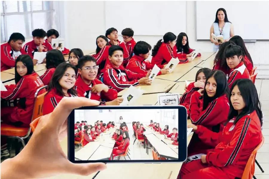 Fotografía de un aula con estudiantes de secundaria en uniforme rojo sentados en mesas; una mano sostiene un celular que graba o fotografía a los estudiantes, ilustrando la consigna 'Graben su video'.