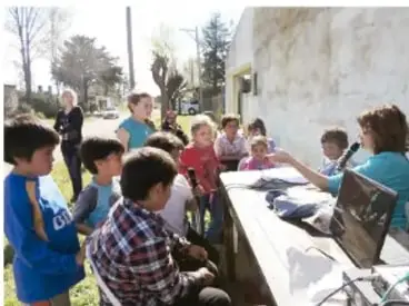 Grupo de estudiantes sentados alrededor de una mesa escuchando a una persona entrevistada, en un espacio al aire libre.