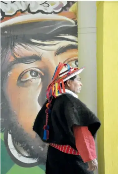 Persona con ropa tradicional (sombrero de plumas y poncho oscuro) que camina frente a un mural de gran formato.