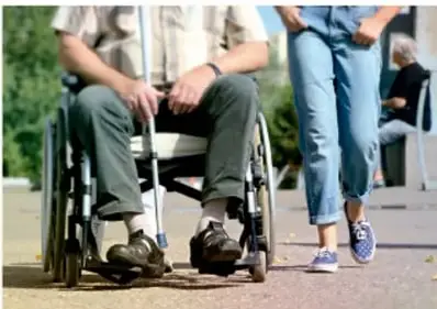Persona en silla de ruedas acompañada de otra persona caminando por una acera.